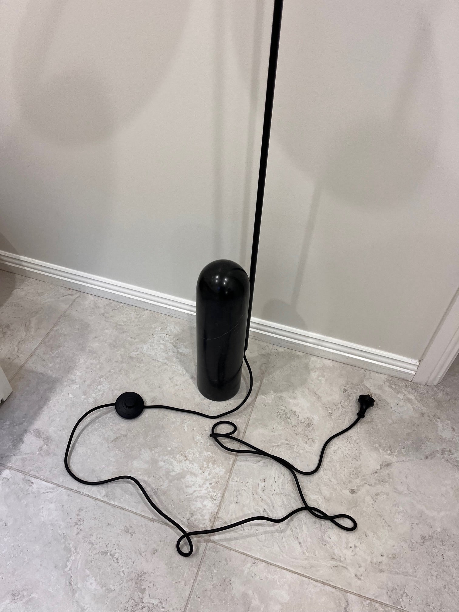 Black cylindrical object with a long black cord on a tiled floor against a white wall.